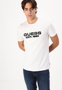 Tricou alb din bumbac cu logo-ul brodat în negru "GUESS EST. 1981" pe față. Mâneci scurte și decolteu rotund. Croială lejeră.