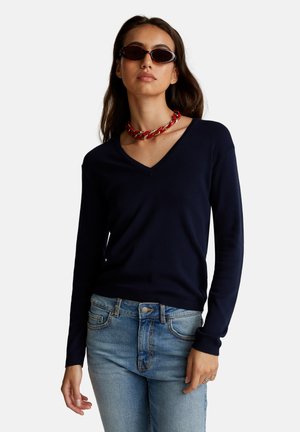 Maglione a V navy in tessuto a costine, design aderente con maniche lunghe, abbinato a jeans azzurri e una collana con catena rossa e dorata.