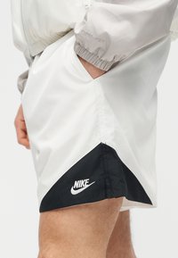 Άτομο που φοράει λευκό και μαύρο σορτς Nike με το χέρι στην τσέπη ανοιχτόχρωμου γκρι μπουφάν, εμφανίζοντας το λογότυπο Nike στο τελείωμα του σορτς.