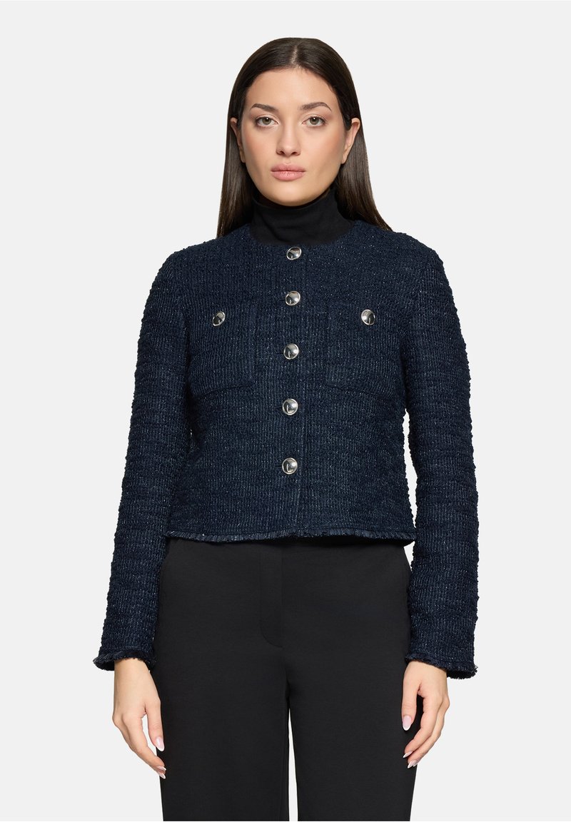 Femme portant une veste boutonnée texturée bleu marine foncé sur un pull à col roulé noir et un pantalon noir, debout devant un fond blanc.