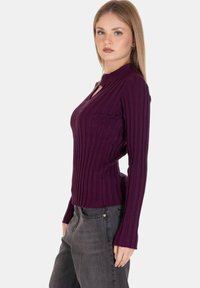 Maglia a maniche lunghe a coste di un luminoso viola scuro con scollatura a chiave e design aderente, indossata con jeans grigi a vita alta. Texture liscia.