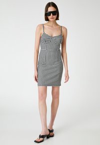 Schwarz-weißes Karokleid mit dünnen Trägern, tailliertem Oberteil und Hüfte, knielangem Saum. Das Model trägt schwarze Sonnenbrillen und High Heels.