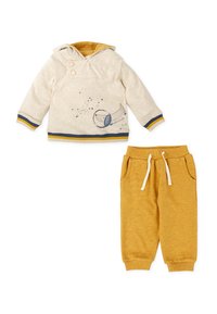 Sudadera con capucha en beige claro con acentos amarillos, que presenta un gráfico de trompeta; combinada con pantalones jogger color mostaza con bolsillos.