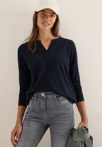 Kvinna som bär en marinblå långärmad skjorta, grå jeans och en beige keps, håller en grön jacka i sin högra hand mot en enfärgad bakgrund.