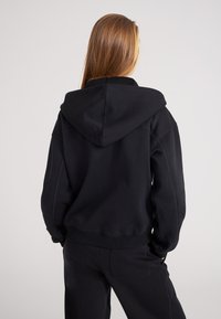 Sudadera negra con capucha y cordón, corte relajado y puños acanalados. Tela suave, detalles de costura mínimos, combinada con pantalones negros a juego.