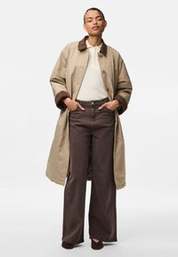 Μπεζ μακρύ παλτό με καφέ σουέτ γιακά και μανσέτες, λευκή ριμπ μπλούζα και σκούρα καφέ φαρδιά τζιν, ολοκληρωμένο με καφέ παπούτσια.