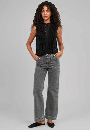 Femme aux cheveux bouclés portant un haut sans manches en dentelle noire, un jean gris taille haute avec jambes larges et des chaussures plates noires, debout les mains dans les poches.