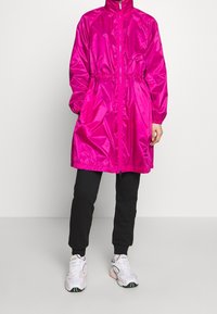 Osoba oblečená ve lesklé jasně růžové dlouhé zipové bundě, černých sportovních kalhotách a bílých sportovních botách na jednobarevném pozadí.