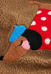 tekstureret brunt stof med en broderet tegneseriehund med en blå hat og en rød polkaprikket frakke. Bemærkelsesværdig brug af levende farver og mønstre.