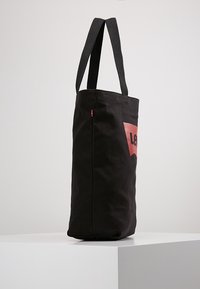 Czarna torba tote z szerokimi paskami z tkaniny. Na froncie widoczny logo Levi's w kolorze czerwonym. Stabilny, wyprostowany kształt.
