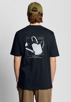 Persoon draagt een zwart T-shirt met een afbeelding van een gieter en een olijfgroene pet, staand tegen een effen grijze achtergrond.