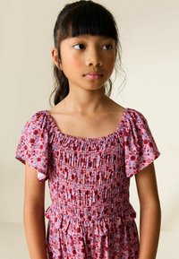 Fiatal lány sötét hajjal, rózsaszín virágos ruhában, ráncolt fűzővel és lebegő ujjakkal, enyhén oldalra nézve, egyszerű háttér előtt.