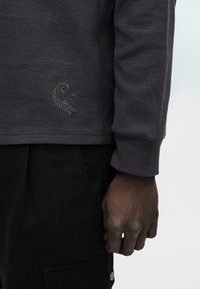 Χέρι με σκούρο χρώμα δέρματος δίπλα στην τσέπη μαύρου cargo παντελονιού, φοράει μακρυμάνικο πουκάμισο σκουρόχρωμης υφής με ένα μικρό κεντητό σχέδιο στροβίλου.