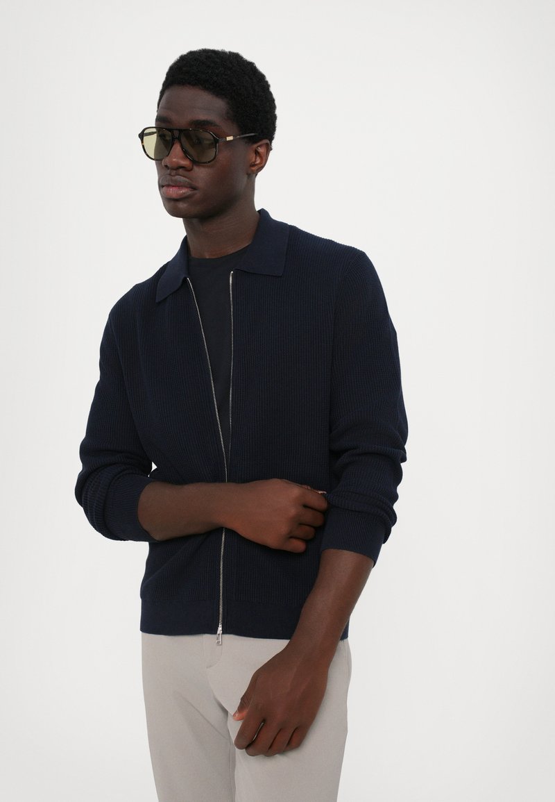 Marineblå ribbet cardigan med lynlås, krave, lange ærmer og struktureret overflade, parret med lys grå bukser.