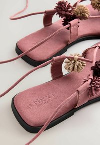 Ružové semišové sandále na plochom podpätku so štvorcovými špičkami, remienkami na obmotanie okolo členka a ozdobnými béžovými a tmavohnedými bambuľkami na remienkoch.