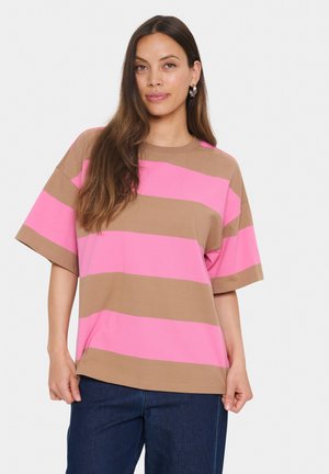 Femme portant une chemise à manches courtes surdimensionnée avec de larges rayures horizontales roses et brunes, associée à un jean bleu foncé, se tenant contre un fond blanc.