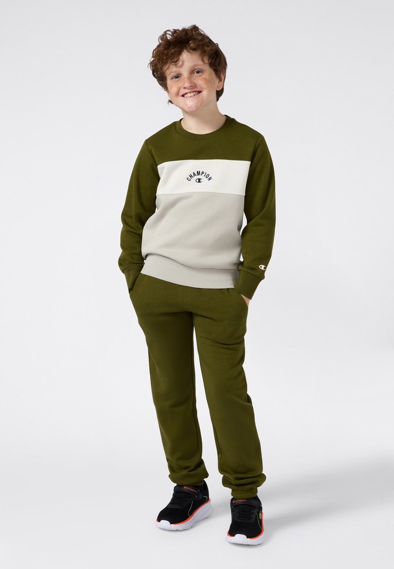 Sudadera verde oliva con franjas crema, logo de "Champion". Joggers a juego en verde oliva y zapatillas negras con acentos rojos. Ajuste cómodo.