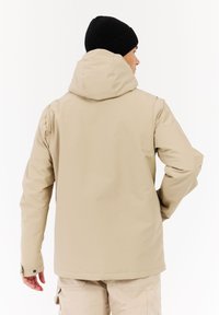 Beige vedenpitävä takki hupulla, joustavilla hihoilla ja sileällä pinnalla. Yhdistetty beigeihin cargo-housuihin, nähtynä takaa.