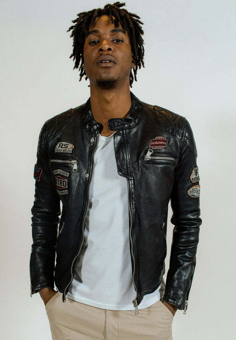 Veste de moto en cuir noir avec des accents matelassés sur les épaules, plusieurs écussons et poches zippées, accompagnée d'une chemise blanche et d'un pantalon beige.