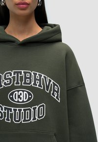 Felpa verde con un grande testo grafico bianco "STUDIO" e "03D" sul davanti. Presenta un cappuccio regolabile con lacci e un tessuto morbido e testurizzato.