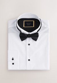 Bílá košile s černými knoflíky, s černým motýlkem a puntíkovaným černým vnitřním límcem. Pečlivě složená, zobrazující detaily manžety.