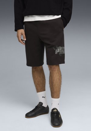 Hombre con pantalones cortos negros Puma con el logo de Puma en la pierna izquierda, calcetines blancos Puma y zapatillas negras Puma sobre fondo gris.