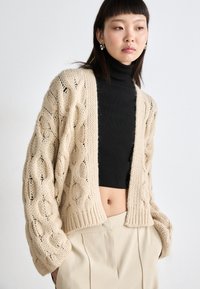 Μπεζ πλεχτό cardigan με καρέ σχέδιο, ανοιχτό μπροστά και υπερμεγέθη μανίκια, φορεμένο πάνω από μαύρη ζακέτα και ανοιχτόχρωμο παντελόνι.