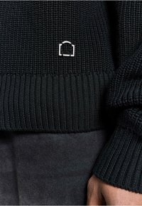 Black ribbed knit sweater featuring a small metallic logo accent at the hem, with a smooth texture and fitted cuffs.