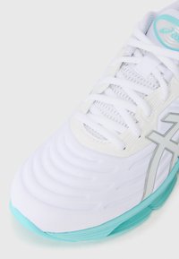 Hvide atletiske sko med en tekstureret overdel, lyseblå sål, sølvdetaljer og snørebåndslukning. Har en mesh-tunge og bølgede design detaljer.