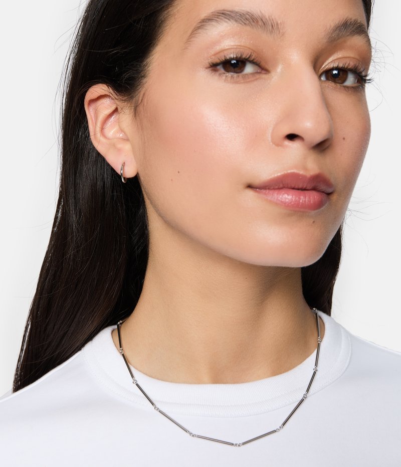 Collana d'argento con link tubular allungati e piccoli accenti rotondi. Indossata su una camicia bianca, evidenziando il suo design elegante.