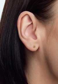 Złota, gwiazdkowata sztyftowa kolczyk na tle koloru skóry, z gładką, błyszczącą powierzchnią i minimalistycznym designem, zapewniający subtelną elegancję.