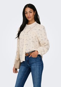 Femme portant un chemisier crème à motif floral avec des détails en dentelle, un jean bleu, une ceinture marron, un sac beige porté à l'épaule, debout devant un fond uni.