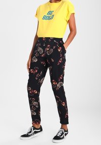 Gul T-shirt med texten "BE BOLD", kombinerad med svarta byxor med ett blommigt paisleymönster. Modellen bär svarta sneakers.