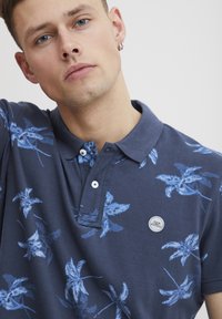 Marineblå polo-skjorte med blått bladmønster, tre-knapp splitt, og brodert logo-merke. Laget av bomull med teksturert overflate.