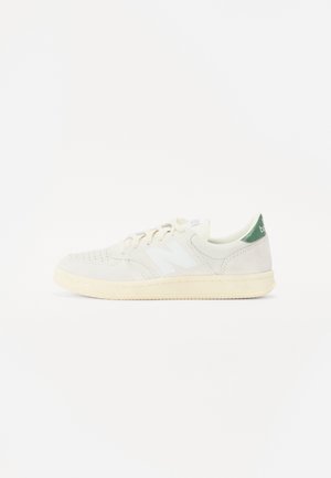 Hvid sneaker med en cremefarvet sål, perforeret tå, hvide snørebånd, et hvidt "N" logo og en grøn hælkappe.