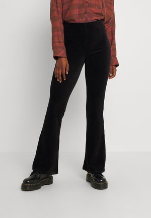 Vrouw die zwarte uitlopende corduroy broek draagt, een rood geruit overhemd en zwarte plateauzolen schoenen, staand tegen een effen witte achtergrond.