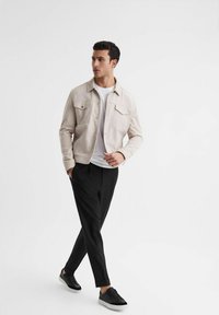 Giacca beige chiaro con due tasche sul petto, indossata sopra una t-shirt bianca, abbinata a pantaloni neri a sigaretta e sneaker nere. Texture lisce.