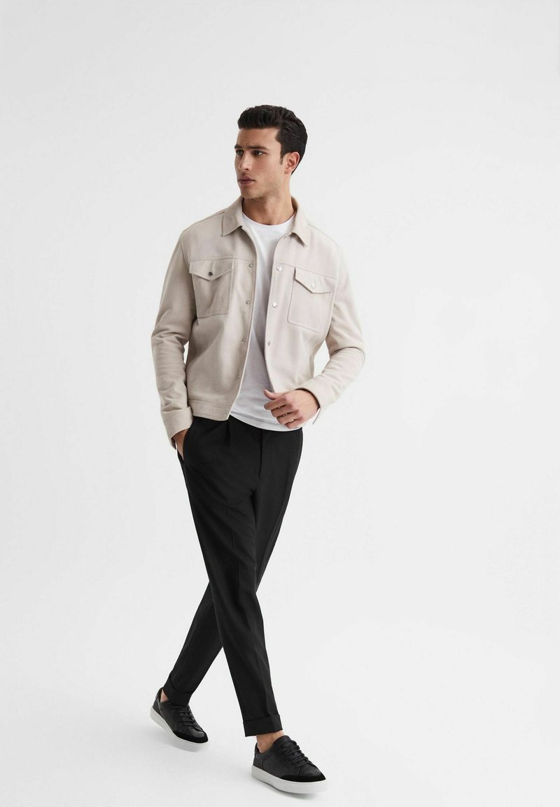 Giacca beige chiaro con due tasche sul petto, indossata sopra una t-shirt bianca, abbinata a pantaloni neri a sigaretta e sneaker nere. Texture lisce.