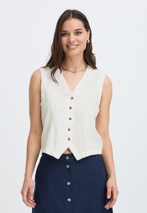 Donna con capelli lunghi e scuri che indossa un top bianco senza maniche con bottoni e una gonna blu scuro con bottoni, sorridente su uno sfondo semplice.