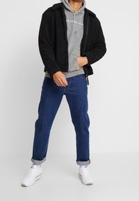 Zwarte fleecejas over een grijze capuchontrui met trekkoord, gecombineerd met blauwe jeans en witte sneakers. Heeft een ontspannen pasvorm en opgerolde manchetten.