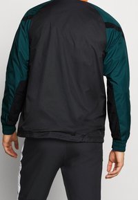 Hombre vistiendo una chaqueta negra y verde oscuro con puños elásticos y pantalones negros con rayas blancas a los lados, visto desde atrás.