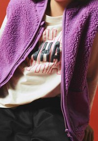 Gilet en polaire violet avec une finition texturée, fermeture éclair et poche latérale, porté sur une chemise de couleur claire ornée de lettres graphiques audacieuses.