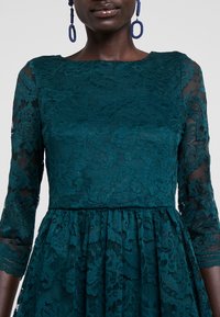 Türkisfarbenes Spitzenkleid mit dreiviertellangen, transparenten Ärmeln, floralen Mustern und einem taillierten Oberteil sowie einem ausgestellten Rock. Strukturierte Stoffdetails sind sichtbar.