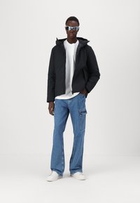 Jachetă padded neagră cu glugă, purtată peste un tricou alb, asociată cu blugi cargo de un albastru deschis și adidași albi. Modelul poartă ochelari de soare cu lentile reflectorizante.