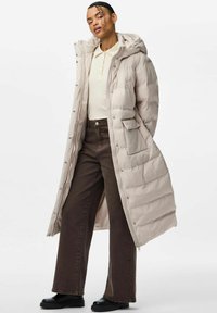 Cappotto lungo e leggero in beige chiaro con cappuccio, chiusura a bottoni a scatto, tasche laterali e texture liscia e trapuntata. Abbinato a pantaloni marrone scuro.