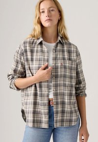 Kariertes Flanellhemd in verschiedenen Beige- und Grautönen, mit Knopfleiste, Brusttasche und aufgerollten Ärmeln. Über einem weißen, gerippten Tanktop getragen.
