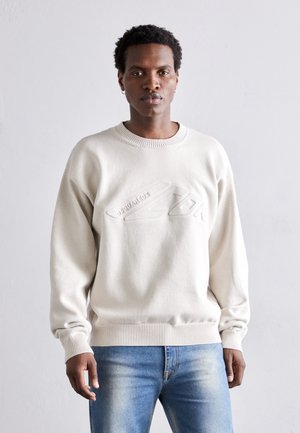 Lys beige sweater med ribbet halsudskærdning og manchetter, med et præget logo på forsiden. Løs pasform, glat tekstur, båret sammen med blå jeans.
