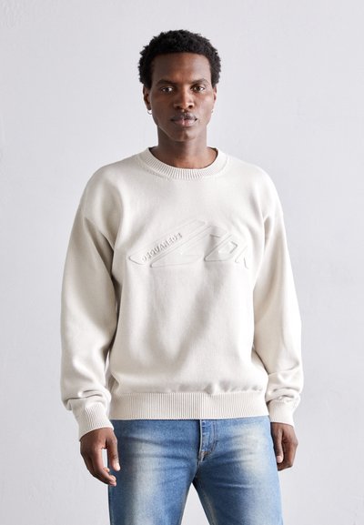 Light beige sweater with ribbed neckline and cuffs, featuring an embossed logo on the front. Casual fit, smooth texture, worn with blue jeans.