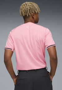 Polo deportivo rosa con detalles negros en las mangas. La tela parece tener textura. Modelo visto desde atrás, con pantalones oscuros.