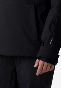 Černá voděodolná bunda s hladkým textilním povrchem, která má manžetu se zapínáním na suchý zip a kapsu na boku.